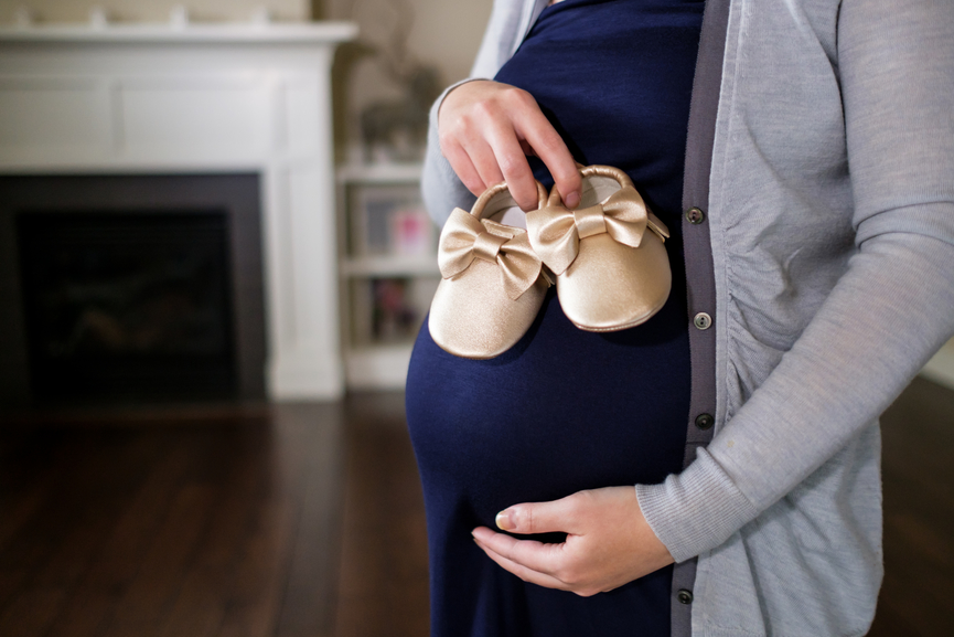 Pregnant Asian woman holding baby shoes at home